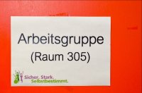 Ein weißes Schild vor einer roten Tür. Auf dem Schild steht: Arbeitsgruppe, Raum 305. Unten auf dem Schild ist ein Logo, es heißt: Sicher. Stark. Selbstbestimmt.